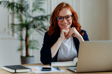 A young woman is sitting in front of a desk smiling into the camera. She is holding a pen. On the desk in front of her we can see books, a mobile phone and a laptop.